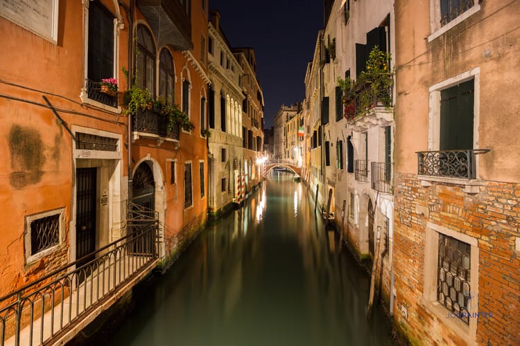 A photograph down a Venetian canal at night. The buildings converge in the distance on a bridge across the canal.